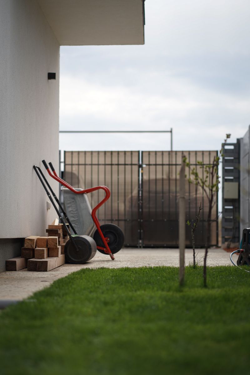 A wheelbarrow and lawn roller beside a house wall, with a green lawn in the foreground